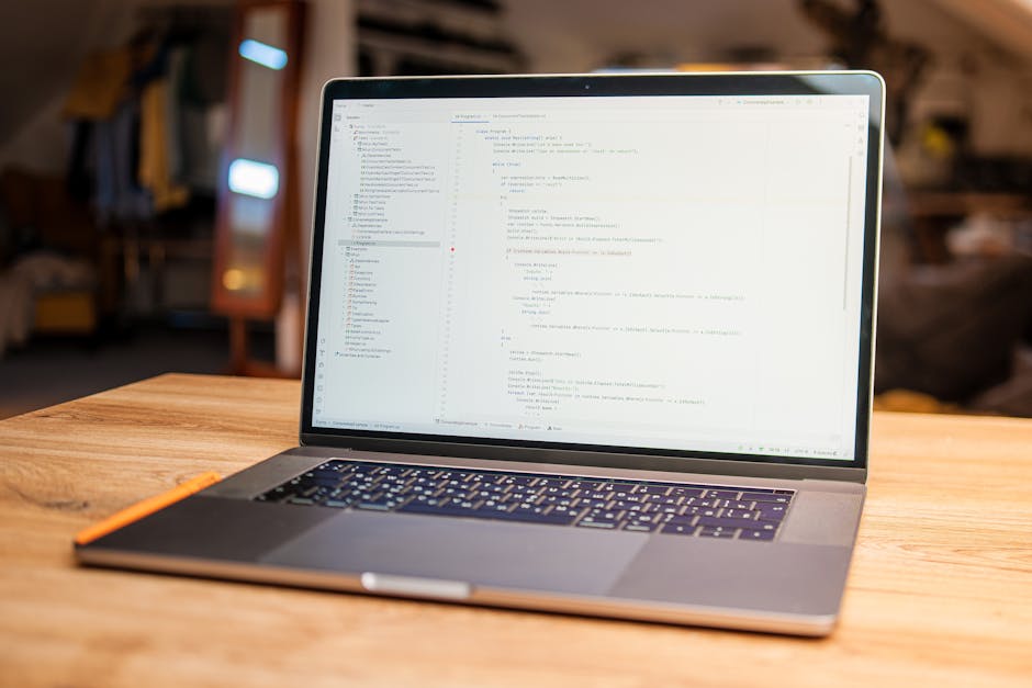 A silver laptop computer with a black keyboard is positioned open on a wooden table, displaying lines of code or text on its screen. The laptop is inside a residential space with a cozy, cluttered bac